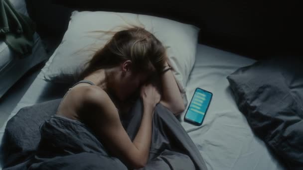 Odgórne ujęcie nerwowej dziewczyny leżącej w łóżku obok telefonu komórkowego z wiadomościami tekstowymi wyświetlanymi na ekranie i zestresowanej przez cybernękanie - Materiał filmowy, wideo