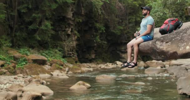 Un viaggiatore solitario siede tranquillamente e riposa su una pietra che pende sull'acqua vicino al fiume sullo sfondo della foresta e del letto del fiume.. - Filmati, video