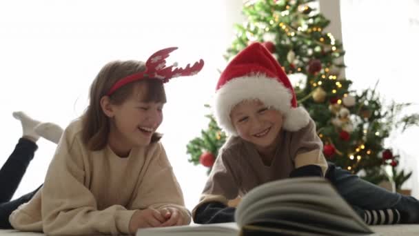 Παιδιά σε ένα καπέλο Σάντα διαβάζοντας ένα βιβλίο και γελώντας, ενώ βρίσκεται στο πάτωμα στο σαλόνι μπροστά από το δέντρο της Πρωτοχρονιάς.  - Πλάνα, βίντεο