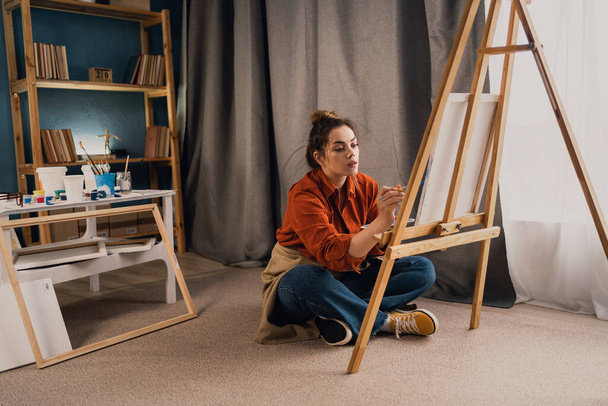 Concentrated Caucasian woman artist sitting on floor in studio painting drawing on canvas, creativity work. Copy space - Photo, Image