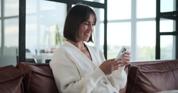 Smiling Caucasian woman relaxes on the sofa while using phone. Her cheerful expression reflects a moment of contentment as she effortlessly navigates device. - Felvétel, videó