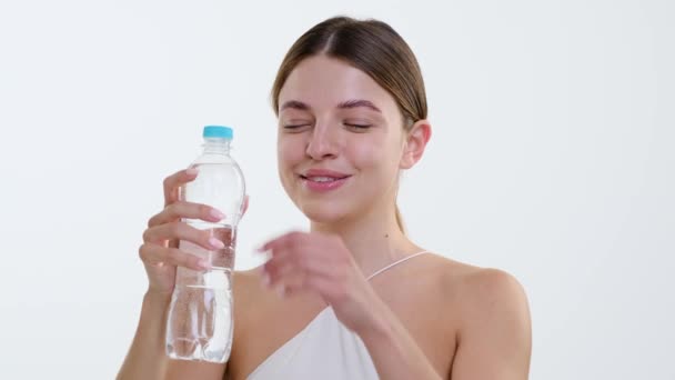 Mulher caucasiana alegre toma um gole refrescante de uma garrafa de água, felicidade evidente no momento da hidratação. Contra o pano de fundo branco limpo, o contentamento brilha através. - Filmagem, Vídeo