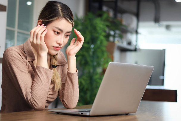 Poważna zmęczona kobieta piękna Azjatka wkładająca głowę w ręce podczas korzystania z laptopa. Przemyślana zagubiona młoda kobieta czuje się zestresowana i ma bóle głowy podczas pracy na komputerze w domu.  - Zdjęcie, obraz