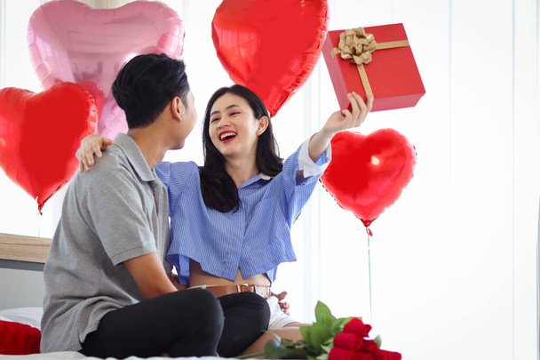 Feliz pareja asiática celebran aniversario juntos, novio dar regalo caja de regalo a la novia sorprendida que necesitan abrazarlo, amante romántico celebrando el día de San Valentín en dormitorio bellamente decorado. - Foto, imagen