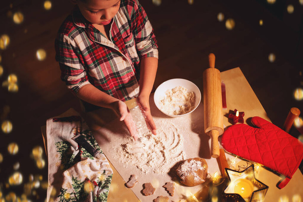 Un petit garçon en pyjama à carreaux rouges sirote de la farine sur la table tout en préparant des biscuits au pain d'épice de Noël. - Photo, image