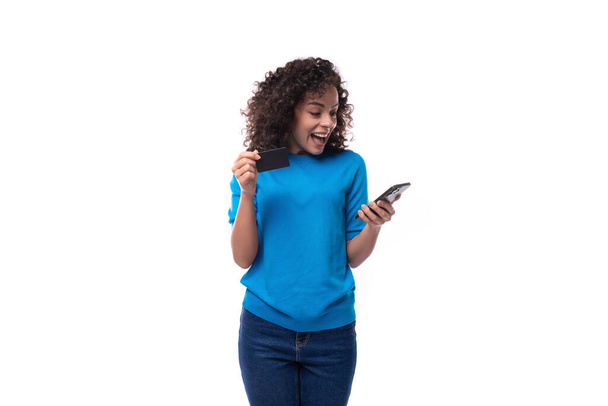 Kıvırcık saçlı genç bir esmer kadın elinde plastik bir banka kartı ve akıllı bir telefonla mavi bir ceket giyiyor.. - Fotoğraf, Görsel