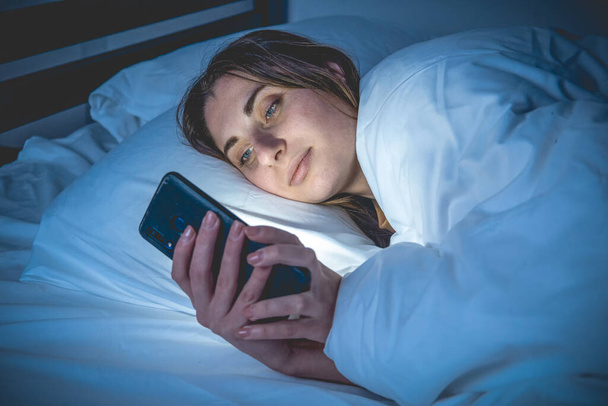Una chica en la cama con un teléfono en una cama blanca en una habitación oscura - Foto, imagen