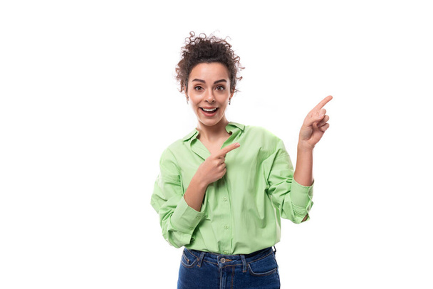 una joven morena rizada modelo femenina delgada vestida con una camisa verde claro señala con su mano al espacio publicitario. Concepto de negocio electrónico. - Foto, imagen