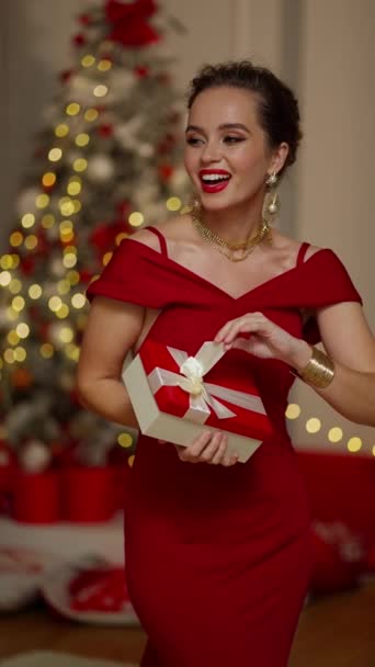 Красивая брюнетка в красном платье рядом с елкой открытая коробка с подарком. - Кадры, видео