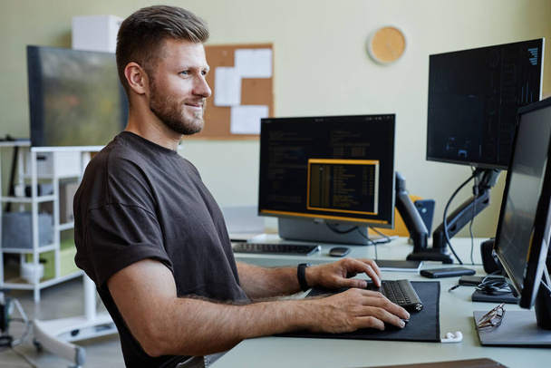 Vue de côté portrait de souriant homme caucasien comme programmeur informatique écriture code au bureau lieu de travail - Photo, image