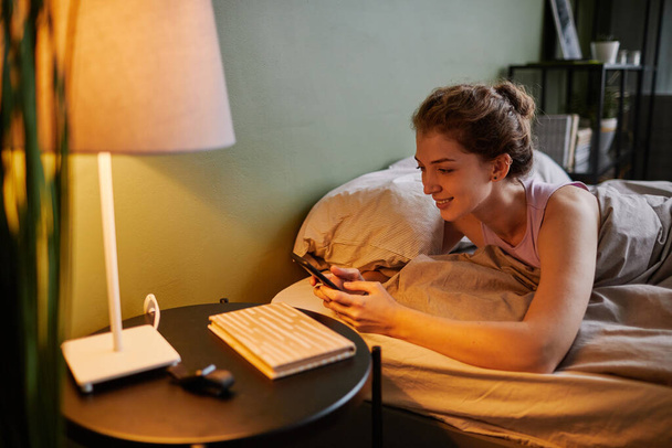Young woman lying on bed in bedroom and communicating online using her smartphone - Photo, Image