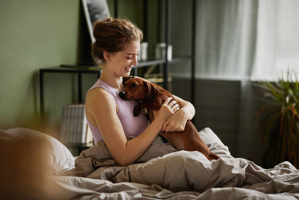 Boldog fiatal lány játszik a kutyájával az ágyban, miközben reggel felébred a hálószobában. - Fotó, kép