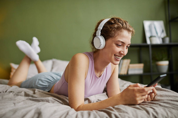 Молодая женщина в беспроводных наушниках отдыхает на кровати и разговаривает в сети со своим смартфоном - Фото, изображение