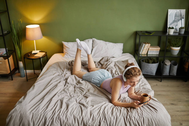 Vista de ángulo alto de la mujer joven en auriculares con teléfono móvil mientras está acostada en su cama con una taza de café - Foto, Imagen