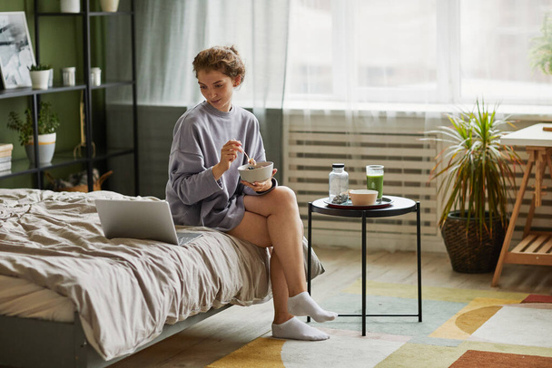 Νεαρή γυναίκα παίρνει πρωινό και χρησιμοποιεί φορητό υπολογιστή, ενώ κάθεται στο κρεβάτι στην κρεβατοκάμαρα στο σπίτι - Φωτογραφία, εικόνα