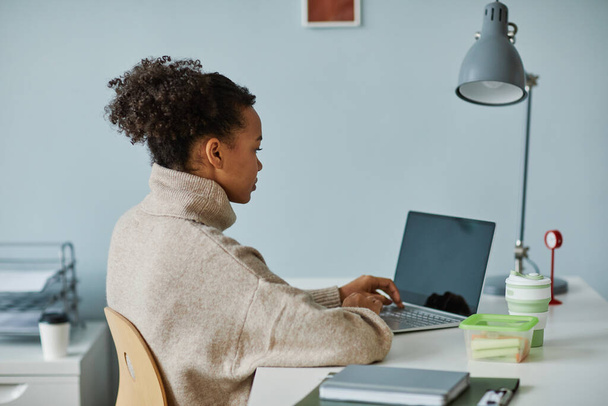 Взгляд на молодую африканскую предпринимательницу, сидящую на ноутбуке на рабочем месте в офисе и выполняющую онлайн-работу - Фото, изображение
