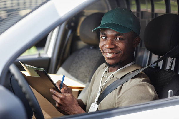 Portrait de jeune homme noir écrivant sur presse-papiers dans la camionnette de livraison et souriant à la caméra, espace de copie - Photo, image