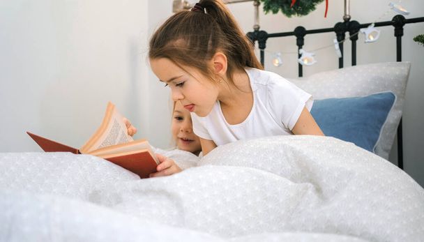Twee zusjes lezen een boek in een bed versierd met kerstverlichting. - Foto, afbeelding