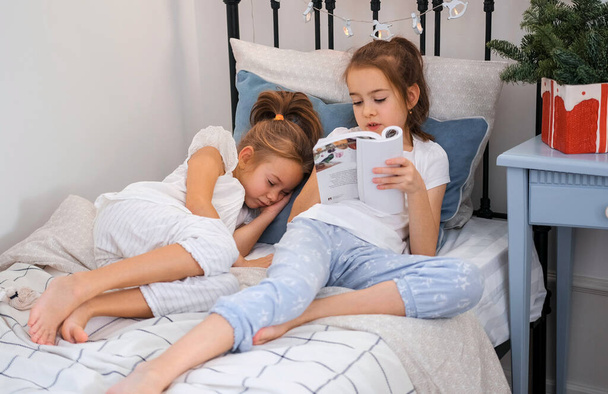 una hermana mayor está leyendo un libro en la cama a su hermana menor que se ha quedado dormida. Noche de Navidad. - Foto, Imagen