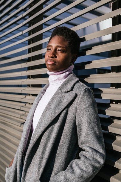 Jonge Afro-Amerikaanse vrouw in stijlvolle jas en trui met kort haar hand in hand en wegkijkend. Ze leunt overdag op een muur van eigentijds gebouw in de stad. - Foto, afbeelding