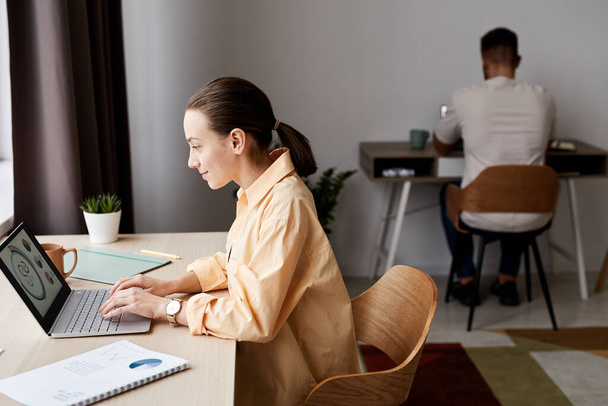 Zijaanzicht van jonge zelfverzekerde brunette zakenvrouw op zoek naar grafische gegevens op laptop scherm tijdens het zitten door werkplek thuis - Foto, afbeelding