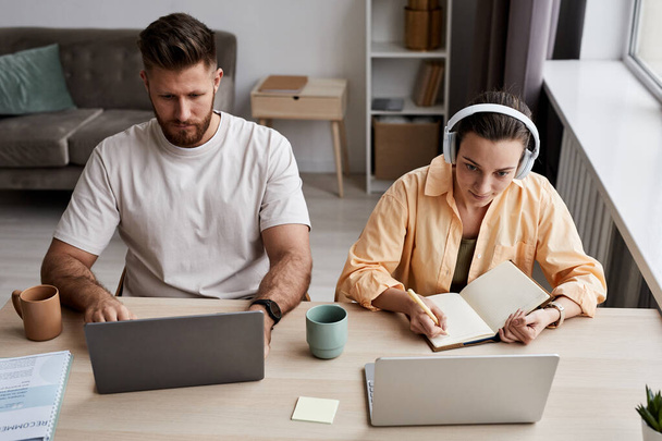 Junge Frau mit Kopfhörer macht sich Notizen im Copybook, während sie während des Online-Unterrichts am Schreibtisch neben einem ernsthaften Kerl vor dem Laptop sitzt - Foto, Bild