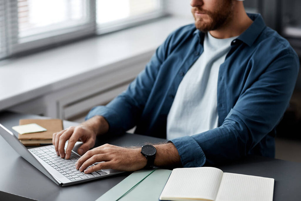 Zbliżenie młodzieńca wpisującego na klawiaturze laptopa siedząc przy biurku i szukając informacji online lub wykonując zadanie - Zdjęcie, obraz