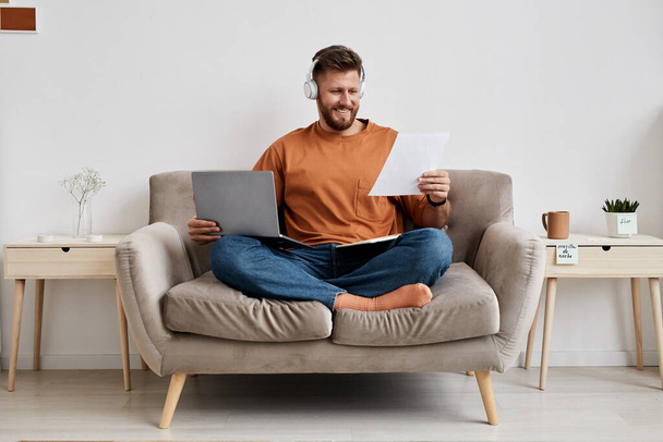 Onnellinen nuori mies, jolla on kannettava tietokone polvillaan katsomassa paperia, jossa on uuden aiheen pisteitä, kun hän istuu sohvalla online-oppitunnin aikana - Valokuva, kuva