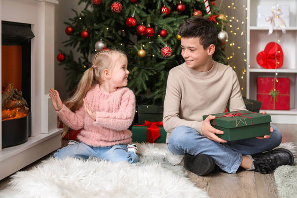 自宅で暖炉の近くに座っているクリスマスプレゼントを持つ小さな子供たち - 写真・画像