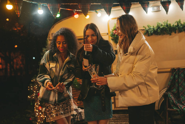 Szczęśliwe dziewczyny świętujące Boże Narodzenie i Nowy Rok sezon zimowy na świeżym powietrzu. Młode, zróżnicowane kobiety radosne przyjaciółki spędzające razem czas dobrze się bawią pijąc wino musujące w pobliżu starej przyczepy xmas - Zdjęcie, obraz