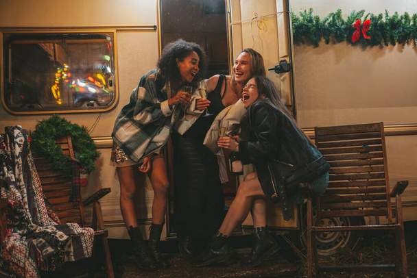 Happy girls celebrating Christmas and New Year winter holidays season outdoor. Young diverse women joyful female friends spending time together have fun drinking sparkling wine near old xmas trailer - Photo, Image