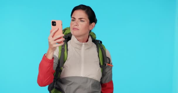 Hiking, phone and a woman in studio for connection, network or search for signal. Young female hiker with a backpack and smartphone for travel, adventure or journey while lost on a blue background. - Footage, Video