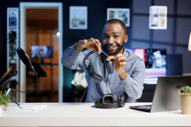 Vlogger afroamericano haciendo video vlog de tecnología, mostrando pares de auriculares recién liberados a los espectadores. Hombre sonriente saludando a los suscriptores, presentando equipo de escucha de música inalámbrica - Foto, imagen