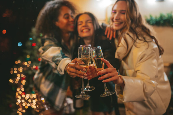 Happy girls celebrating Christmas and New Year winter holidays outdoor. Молодые разнообразные женщины радостные подруги проводить время вместе весело пить игристое вино возле старого трейлера Рождество - Фото, изображение