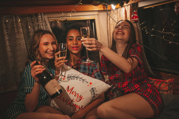 Happy girls celebrating Christmas and New Year winter holidays season in camper. Active young diverse women joyful spending time together hugging have fun drinking sparkling wine in old xmas trailer - Photo, Image