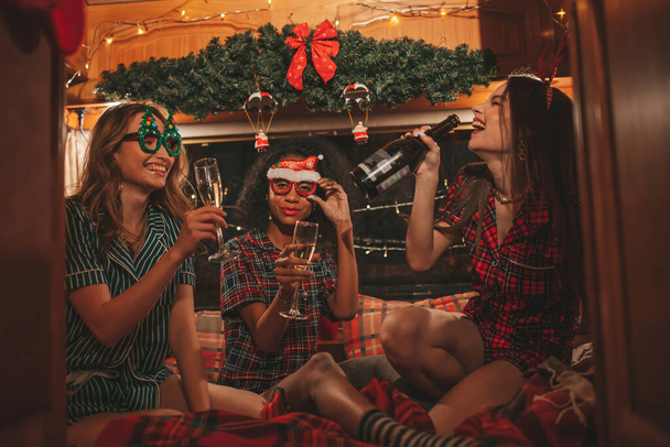 Happy girls celebrating Christmas and New Year winter holidays season in camper. Active young diverse women joyful spending time together hugging have fun drinking sparkling wine in old xmas trailer - Photo, Image
