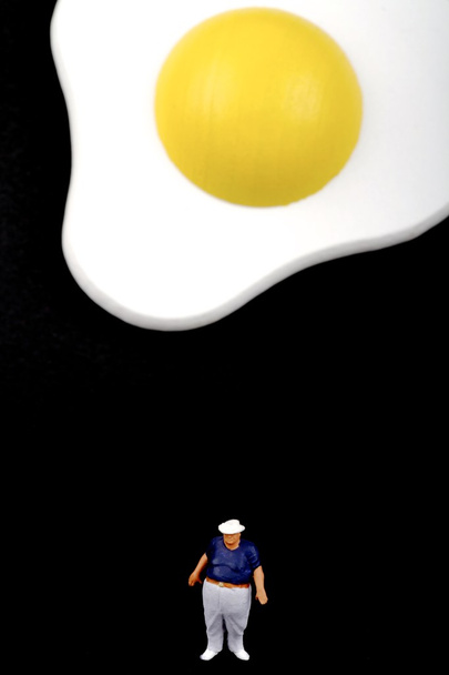 Miniatura de um homem gordo perto por um ovo frito
 - Foto, Imagem