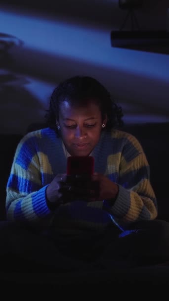 Vertikale Junge schwarze Mädchen, die nachts im Wohnzimmer auf dem Smartphone chatten. Konzentriert und technologiesüchtig sitzt er im Dunkeln gemütlich auf dem Sofa zu Hause. Rollende Social-Media-Generation z. - Filmmaterial, Video