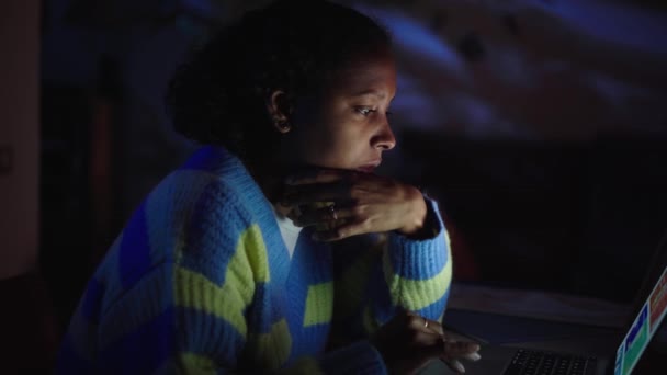 Boční pohled koncentrované mladé latinské dívka pomocí notebooku doma v noci. Vážná žena hledající a píšící na počítači. Lidi sedí v obýváku. Černé ženské osoby osvětlené obrazovkou zařízení. - Záběry, video