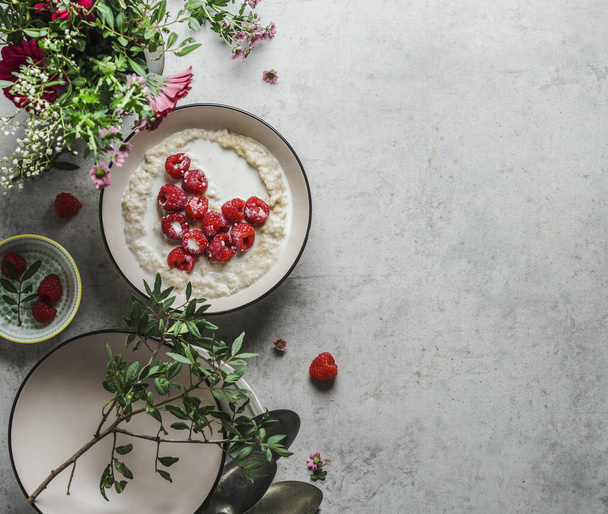 Romantic breakfast with porridge and heart of raspberries on grey kitchen table with flowers and bowls. Vegan breakfast idea with nutritious ingredients. Top view with copy space. - Photo, Image