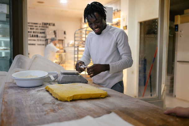Junger schwarzer Bäcker mit Dreadlocks und weißer Kleidung schneidet mit einem Schulterblatt gelben Teig und wiegt ihn in einer Bäckerei auf einer Waage - Foto, Bild