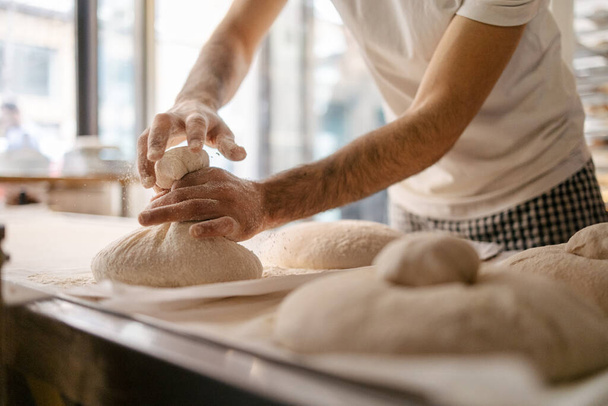 Руки невпізнаваного чоловічого пекаря в білій футболці, що захоплює хлібне тісто і формує його перед тим, як покласти його в духовку в пекарню - Фото, зображення