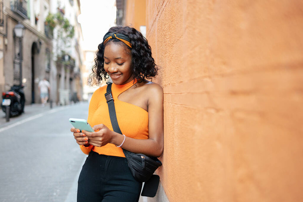 Πορτρέτο μιας όμορφης νεαρής μαύρης γυναίκας που φοράει καθημερινά ρούχα χρησιμοποιώντας το smartphone του χαμογελώντας ενώ ακουμπά σε έναν πορτοκαλί τοίχο - Φωτογραφία, εικόνα
