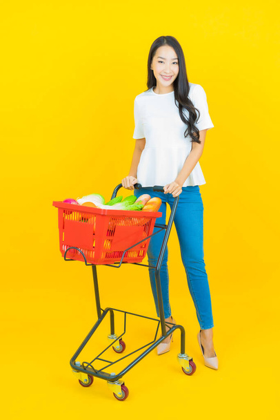 Portrét krásná mladá asijská žena úsměv s nákupním košíkem ze supermarketu na žlutém pozadí - Fotografie, Obrázek