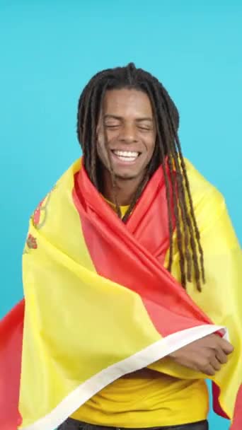 Felice uomo latino con dreadlocks avvolgente con una bandiera nazionale spagnola in studio con sfondo blu - Filmati, video