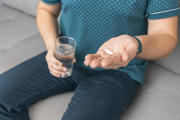Man taking medicine, holding in a hand white therapeutic pills, antibiotics, painkiller and glass of water, close-up view. - Photo, Image