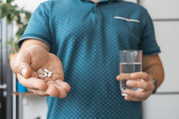 Mężczyzna bierze leki, trzyma w ręce różne tabletki lecznicze, antybiotyki, środki przeciwbólowe i szklankę wody, widok z bliska. - Zdjęcie, obraz