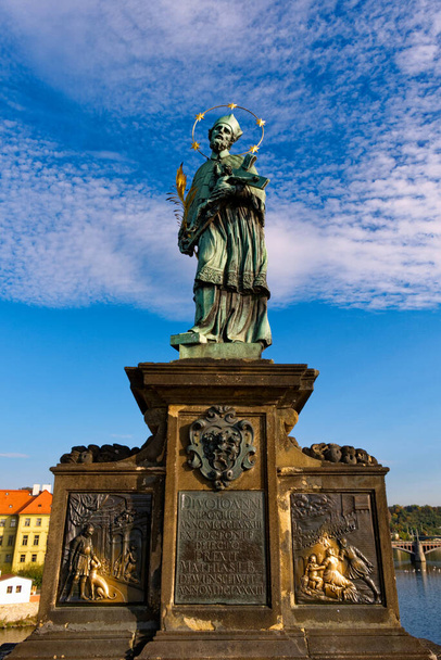 Una statua di un santo con una croce e un libro su un piedistallo. Statua di Giovanni Nepomuceno sul Ponte Carlo a Praga contro il cielo. - Foto, immagini