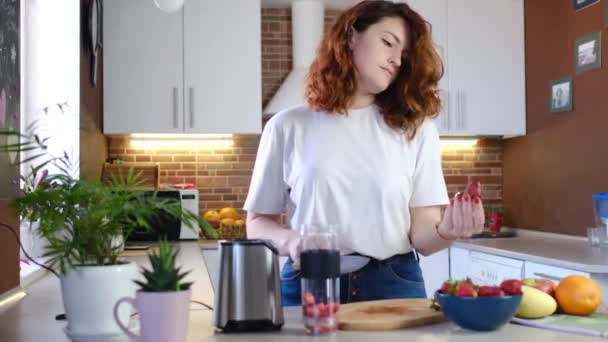 Młoda kobieta w domu w kuchni zrobić koktajl owocowy. Piękna młoda, szczęśliwa kobieta w domowej kuchni przygotowuje koktajl fitness w domu. Mączka sportowa i wegańska - Materiał filmowy, wideo