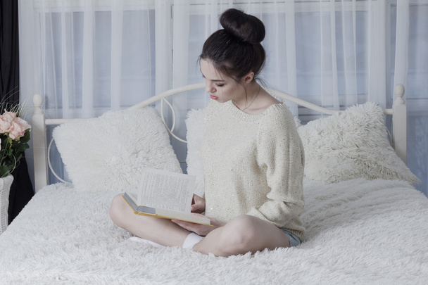 Meisje, zittend op het bed thuis en bladeren door een boek - Foto, afbeelding